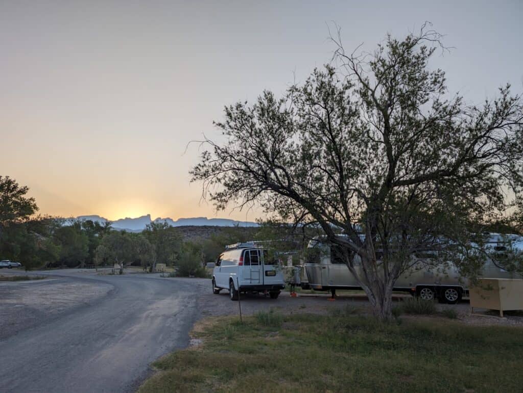 A scenic campground at sunset with shaded RV trailers, a quiet dirt road, and a tree in the foreground, offering a peaceful outdoor adventure experience for campers and outdoor enthusiasts.