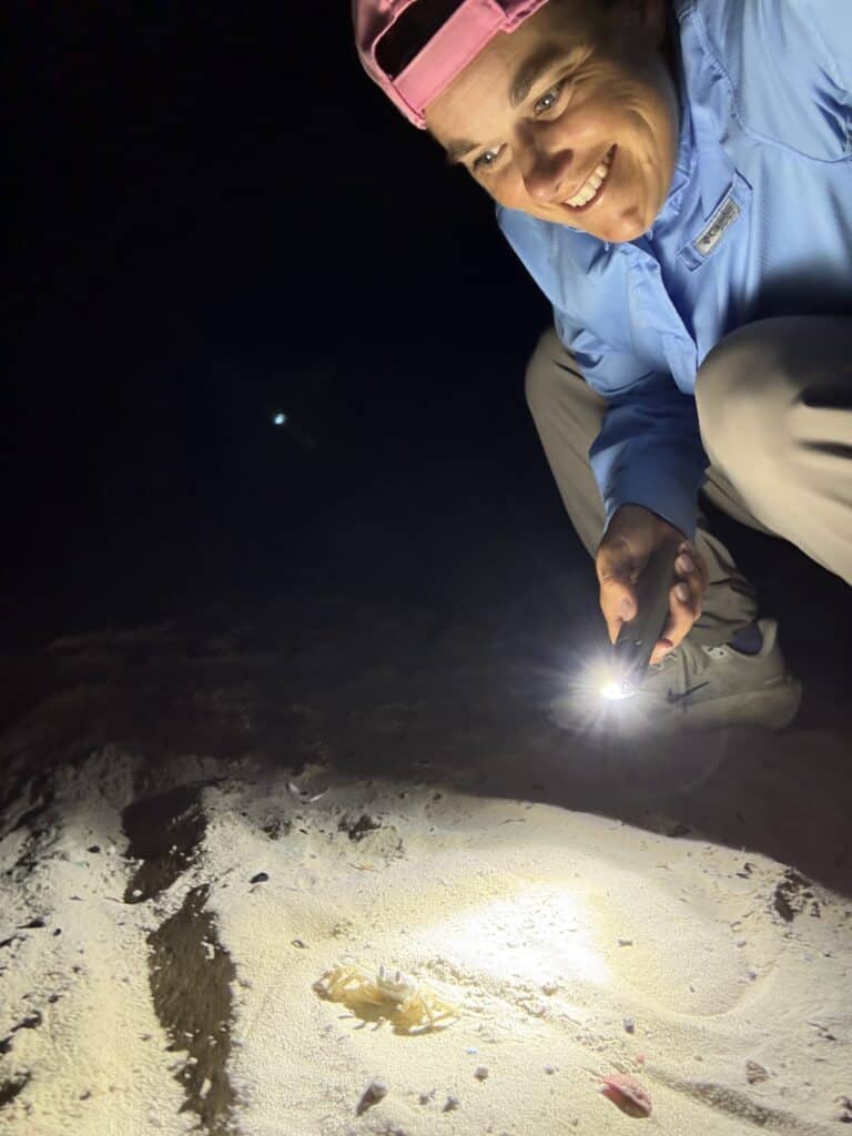 Bright night beach walk with a smiling man holding a flashlight, discovering a crab in the sand, in a serene coastal setting under the stars.