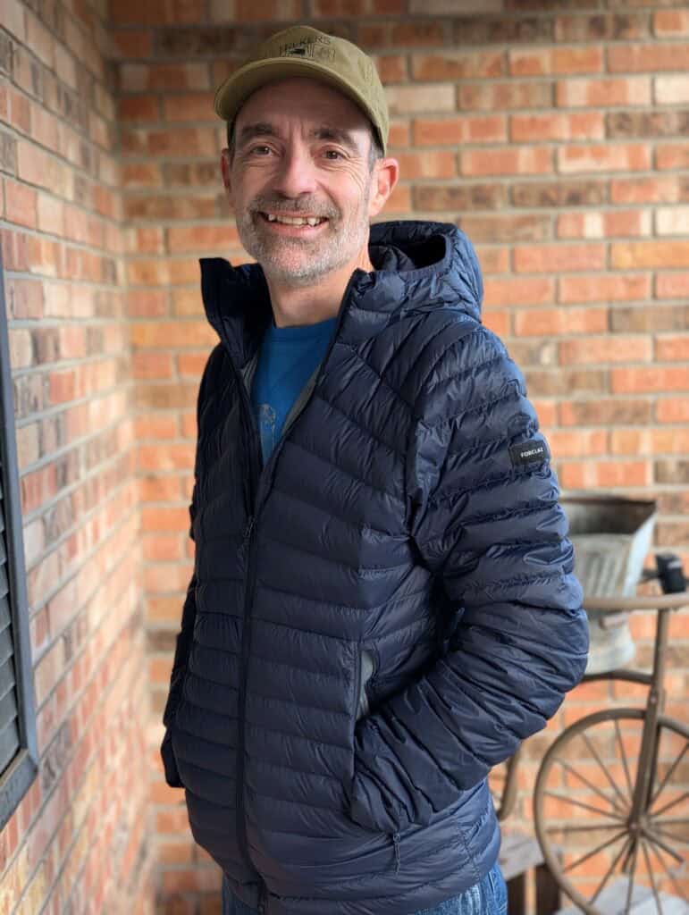 Adventure enthusiast wearing a navy puffer jacket and cap with TREKERS logo, standing against a brick wall, showcasing outdoor gear perfect for trekking and hiking excursions.