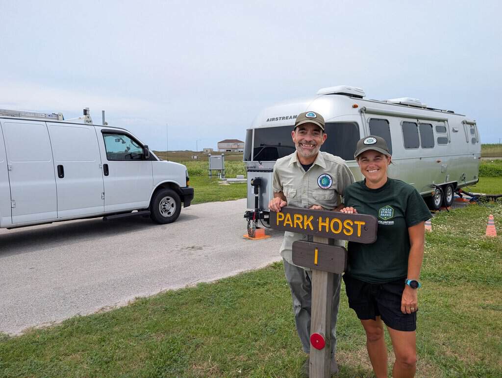 Two park hosts standing in front of a camper trailer and a white van at a campground, smiling and holding a park host sign.